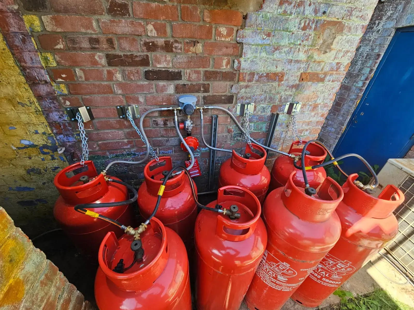 Bank of red LPG cylinders connected to a wall-mounted changeover manifold outside a brick building