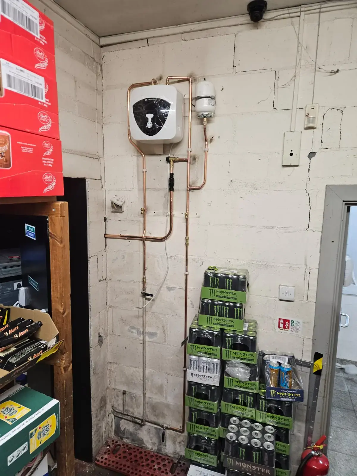 Small undersink-style electric water heater with copper pipework on a shop's back-room wall